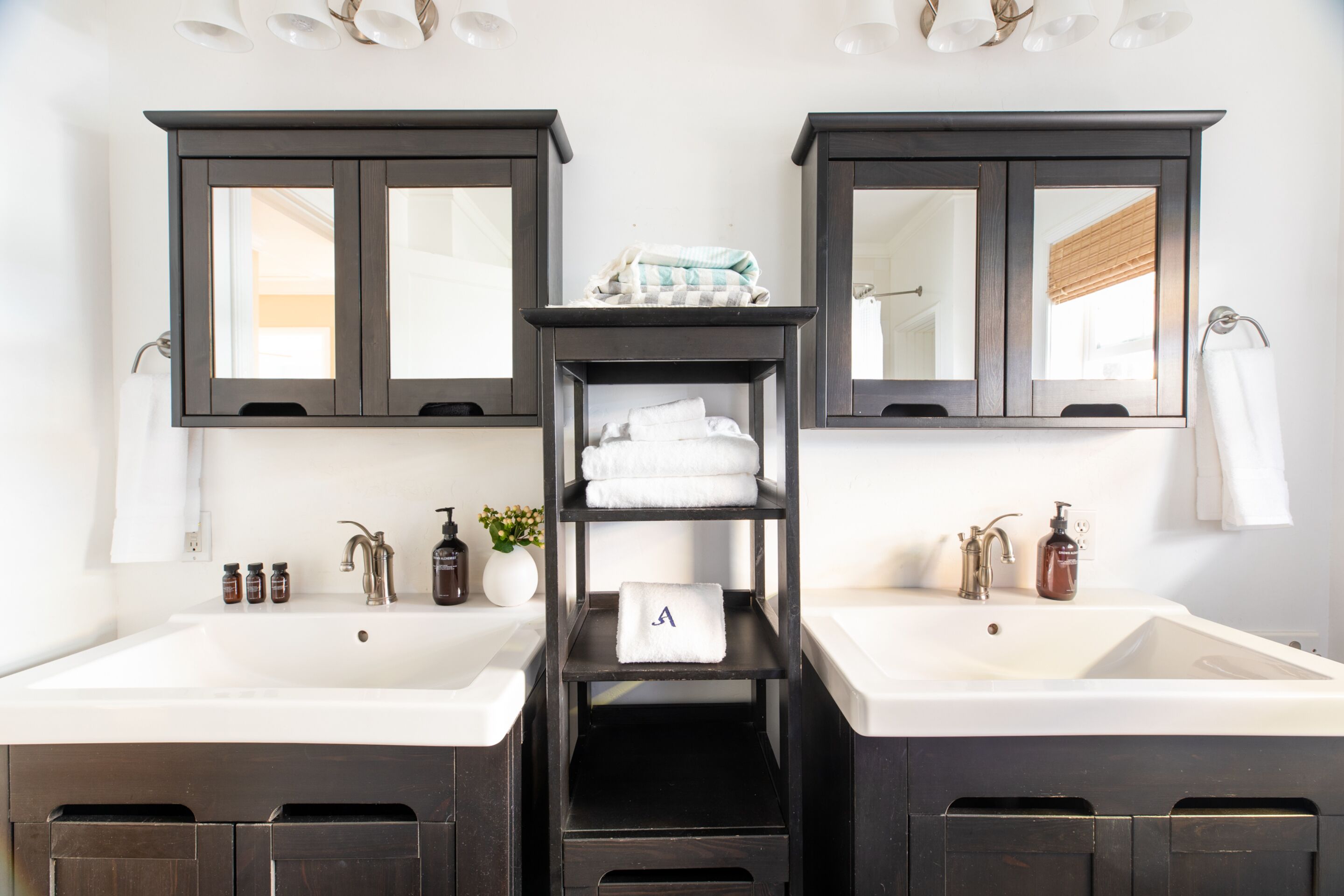 A bathroom with two sinks, dark wooden cabinets, and a central shelf with towels.