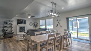 A spacious living room with a fireplace, a dining area with a wooden table and chairs, and a large window with blinds. - Boat Harbor House - Pool & Hot Tub Access in Unbeatable Location! (Garden City)