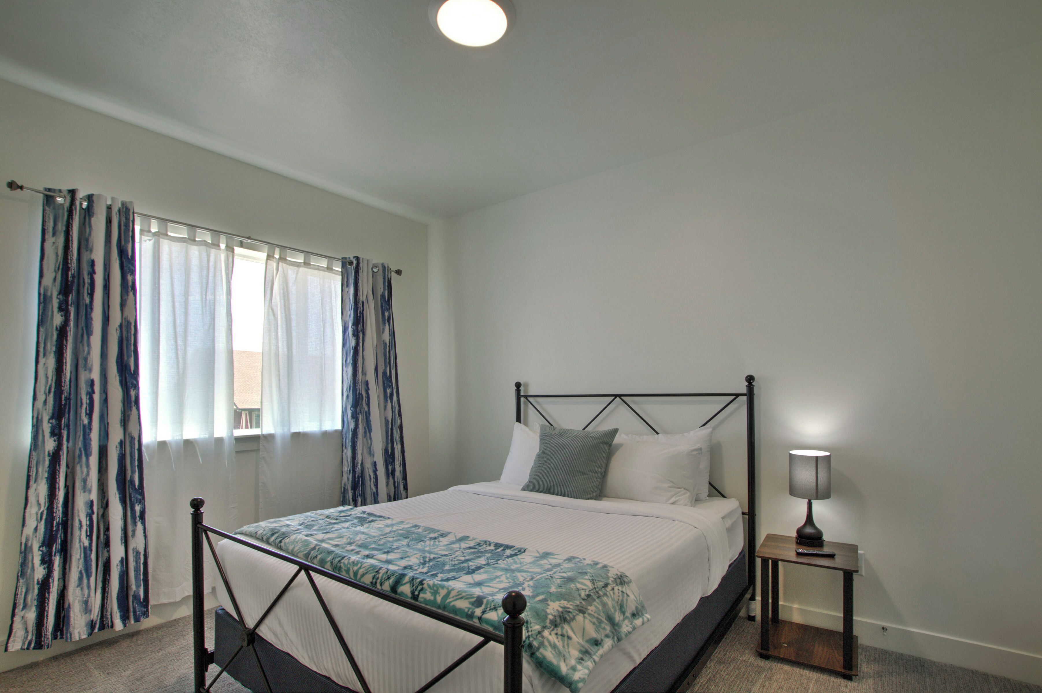 A neatly made bed with a metal frame, a bedside table with a lamp, and a window with curtains.