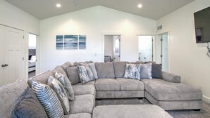 A spacious living room with a large sectional sofa, a flat-screen TV mounted on the wall, and a door leading to another room. - Boat Harbor House - Pool & Hot Tub Access in Unbeatable Location! (Garden City)