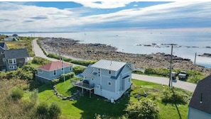Une maison située sur la côte, dotée d’un toit en pente, d’un porche à l’avant et offrant une vue sur le littoral rocheux et la mer.