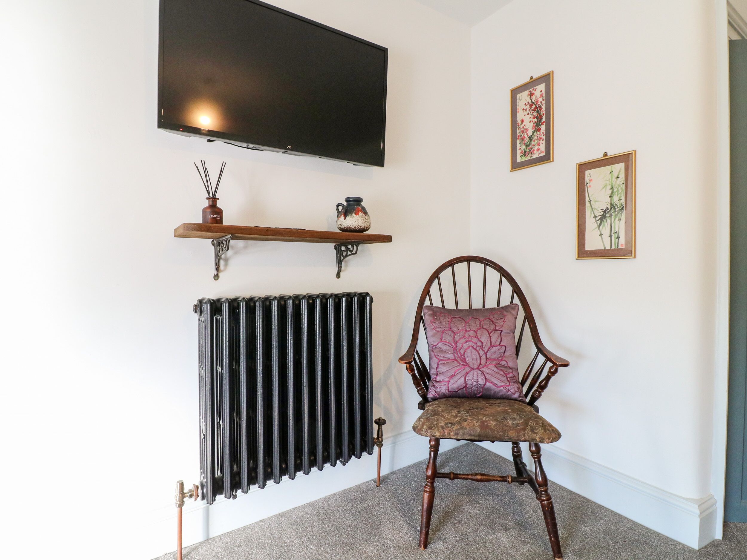A room with a radiator, a chair with a floral cushion, a shelf with decorative items, and a mounted television.