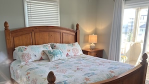 A bedroom with a wooden bed, a ceiling fan, a window with blinds, and a nightstand with a lamp. - Completely Renovated in 2022! Perfect Location! (Kill Devil Hills)