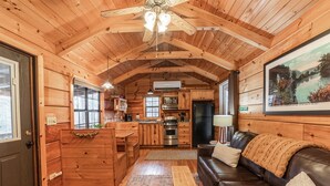 A cozy cabin interior with wooden walls, a kitchen area, a leather sofa, and a ceiling fan. - Wooded Couples Log Cabin w/ Hot Tub (Pet-friendly) (Hot Springs)