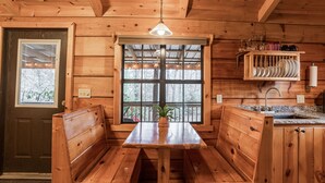 A wooden cabin interior with a central wooden table and bench seating, a window with a view of trees, and a shelf with dishes and glasses. - Wooded Couples Log Cabin w/ Hot Tub (Pet-friendly) (Hot Springs)