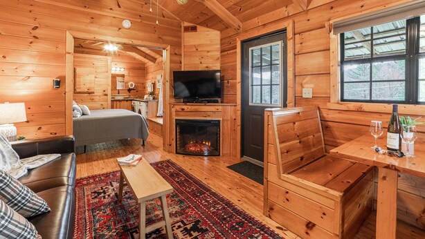 A cozy log cabin interior with a fireplace, wooden furniture, a television, and a large window. - Pet-Friendly Mountain Log Cabin for Couples (Hot Springs)