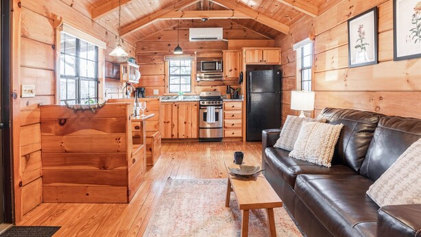 A cozy cabin interior with wooden walls, a kitchenette, a dining area, and a living room with a leather sofa.