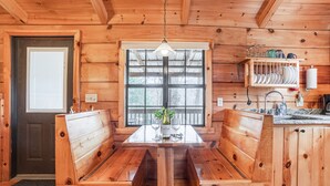 A wooden interior with a central dining table, benches, a window, and a kitchen area with shelves.