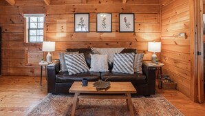 A cozy cabin interior with a wooden ceiling, a leather sofa with patterned cushions, a coffee table, and framed botanical prints on the wall.