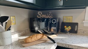 A kitchen counter with a loaf of bread, a knife, a cutting board, a microwave, and a bowl of fruit. - Charming Ranch style house across from Mercyhurst University (Erie)