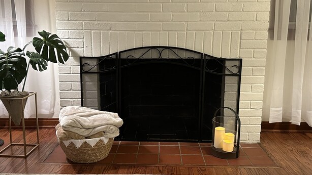A fireplace with a white brick surround, a black metal screen, and a basket of folded blankets. - Charming Ranch style house across from Mercyhurst University (Erie)