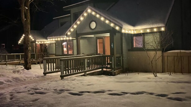 Une maison dotée d’un porche, illuminé par des guirlandes lumineuses, et entourée de neige.