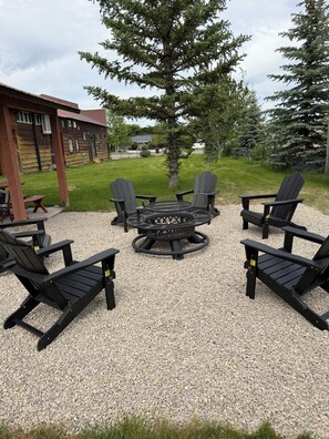 A gravel patio with four Adirondack chairs, a circular fire pit, and a wooden building in the background.