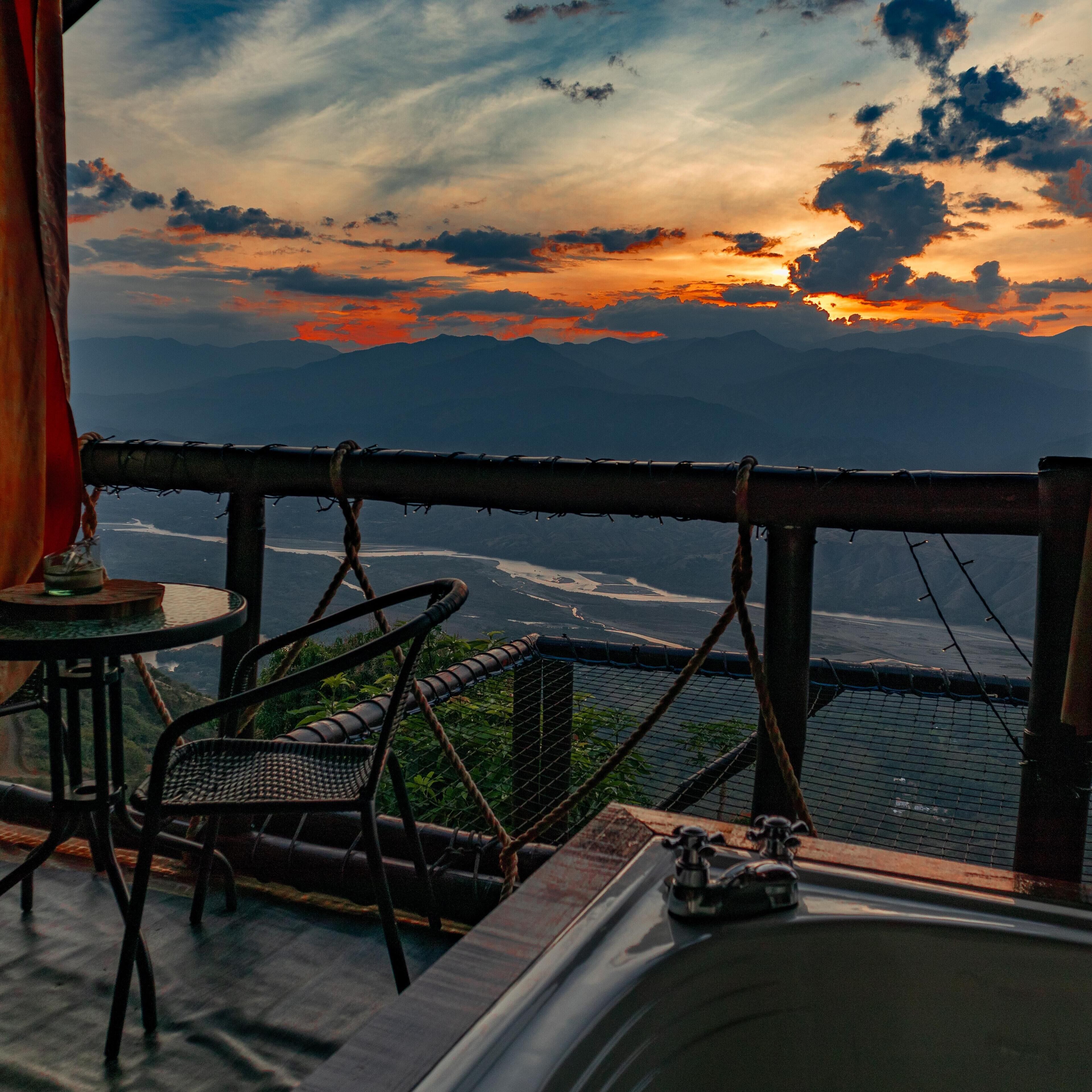 A terrace with a view of a mountainous landscape during sunset. There are wicker chairs and a small table with a glass.