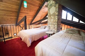 Cozy attic bedroom with sloped ceiling, wooden beams, and stone accent wall, featuring two beds with white bedding and a small table.