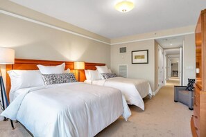 Een hotelkamer met twee bedden, een houten hoofdbord, een bedlampje, een lijst met een afbeelding aan de muur en een gang naar een andere kamer.