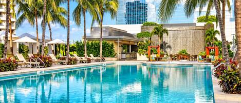 A pool area with lounge chairs and palm trees, surrounded by a modern building with a glass facade.