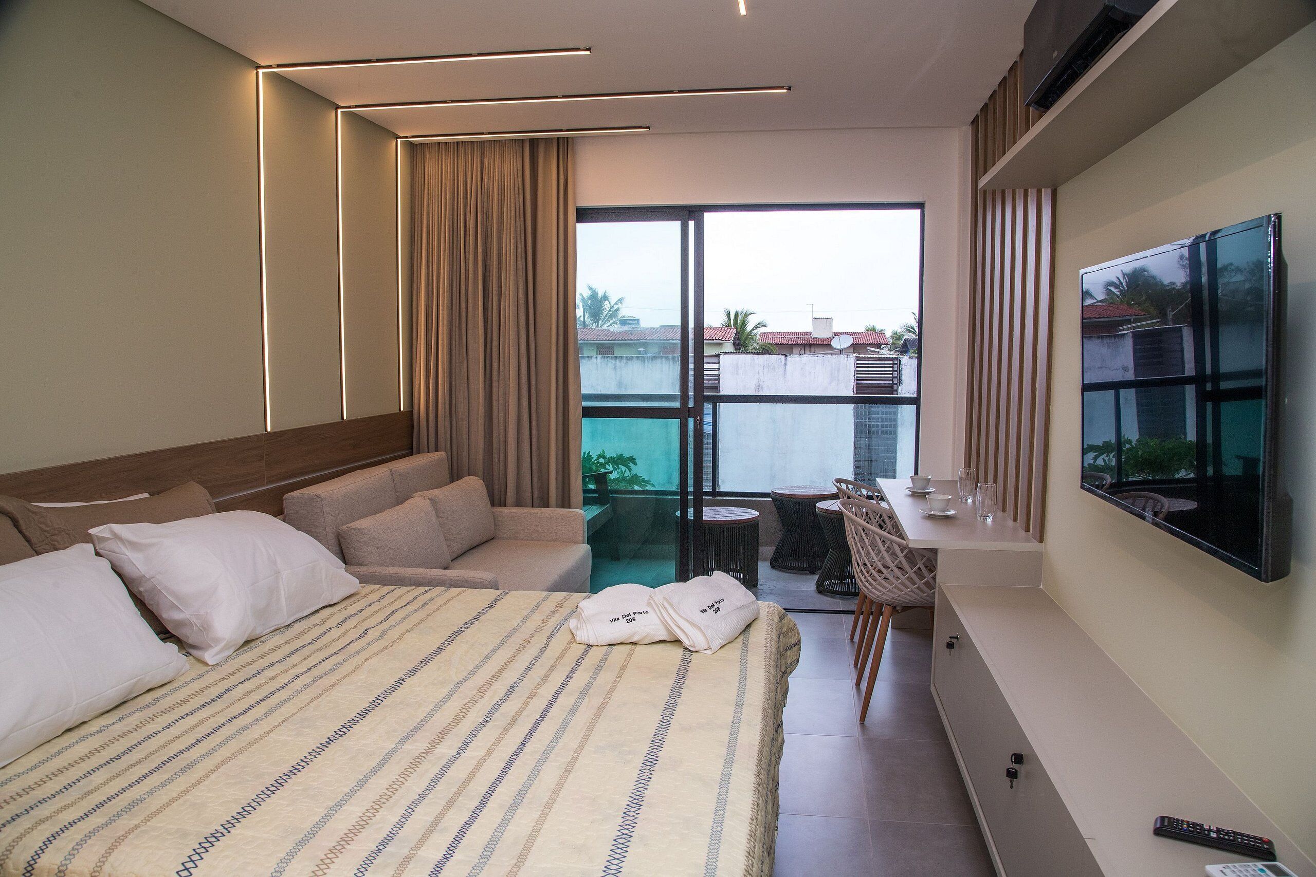 A modern hotel room with a large bed, a sofa, a balcony with a table and chairs, and a TV mounted on the wall.