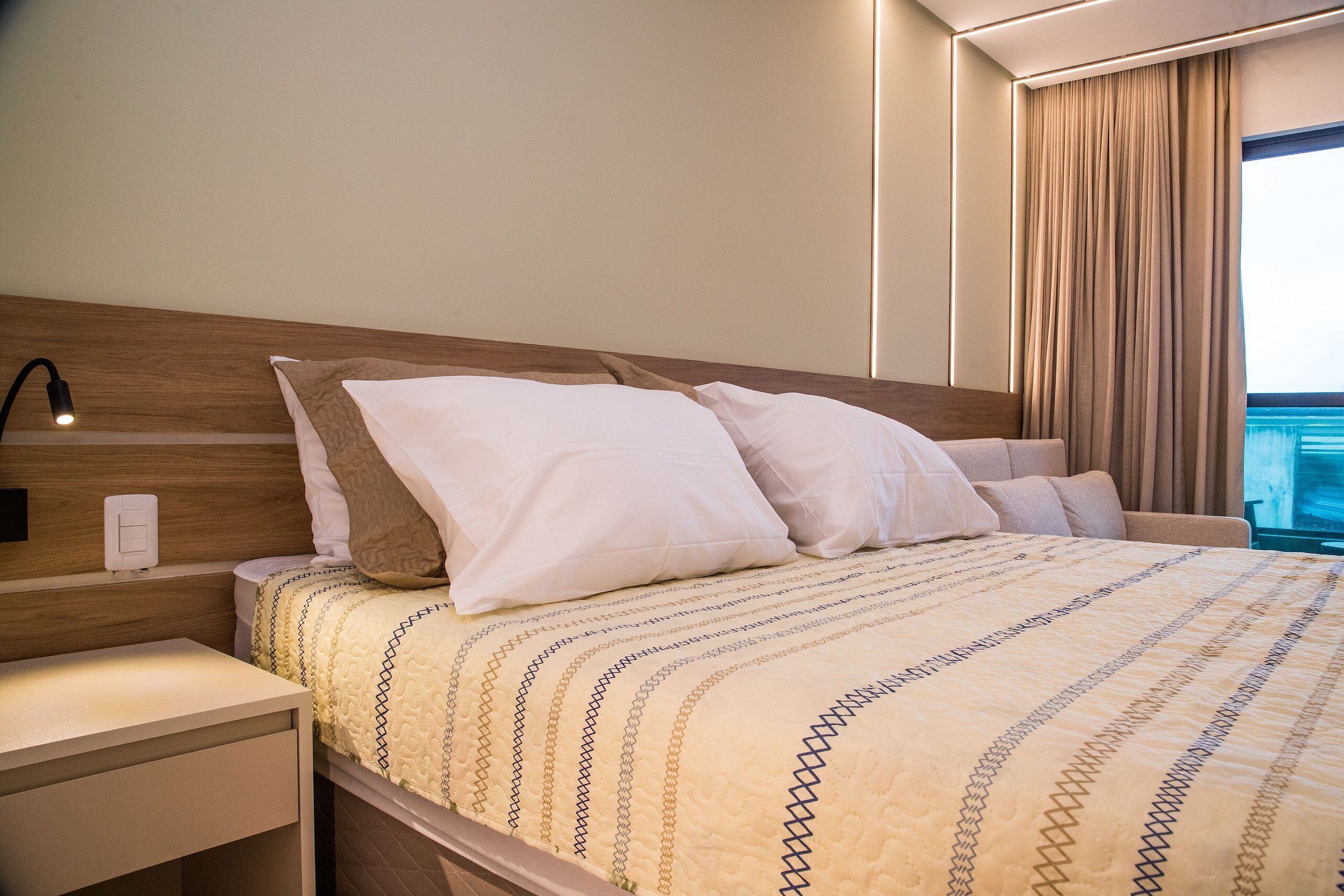 A neatly made bed with a patterned bedspread, a bedside table with a lamp, and a view of the ocean through a window with curtains.