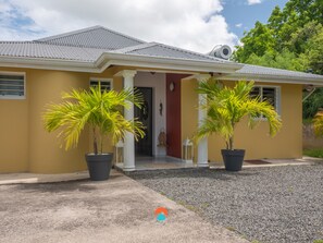 Une maison à la façade jaune, au toit gris et ornée de deux palmiers en pot devant l’entrée.