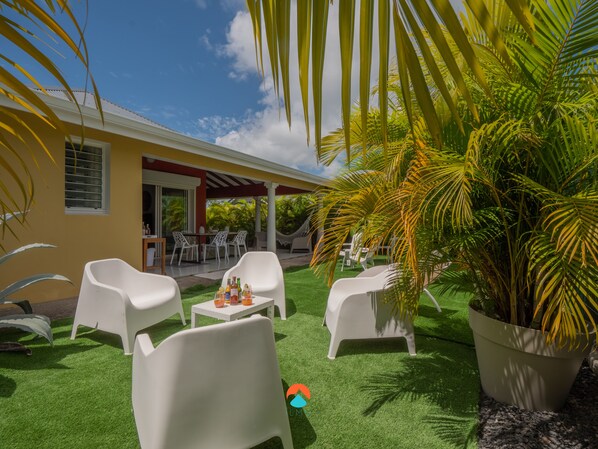 A patio area with white plastic chairs and a small table, surrounded by lush green artificial grass and palm plants.