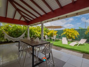 A covered patio with a table and chairs, a hammock, and a view of a lush garden.