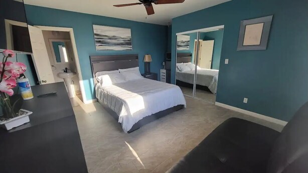 A bedroom with a bed, a desk, a chair, a ceiling fan, and a bathroom with a sink and mirror. - Tranquilo y Comodo. (Compton)