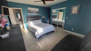 A bedroom with a bed, a desk, a chair, a ceiling fan, and a bathroom with a sink and mirror.