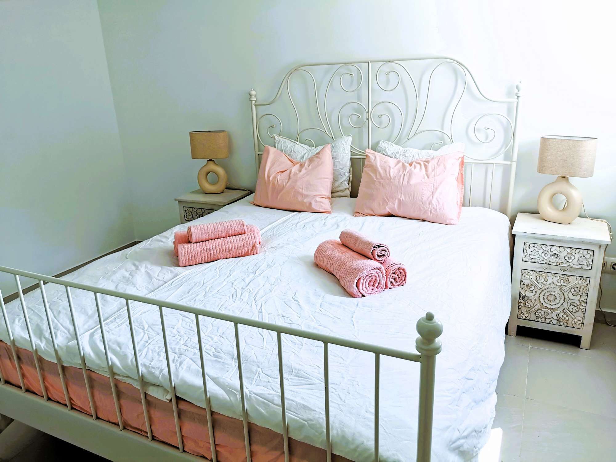 A neatly made bed with pink towels, a white metal headboard, and matching nightstands with lamps.