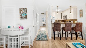 A bright, white interior with a kitchen and dining area. There are wicker chairs, a white table, and a blue rug. - ⭐350 yards to Beach~First Floor~Community Pool~1BR Sandprints A3⭐ (Miramar Beach)
