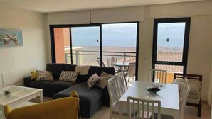 A modern living room with a sofa, a dining table, and chairs. There are large windows offering a view of the cityscape. - Apartment La Verita, Caleta de Fuste (Castillo Caleta de Fuste)