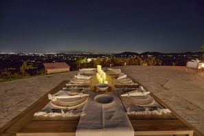 Uma longa mesa de jantar posta para uma refeição com velas, em um terraço com vista para a cidade à noite.