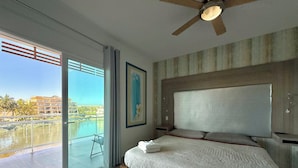 A modern bedroom with a large bed, a ceiling fan, and a view of a body of water and buildings. - Pacifika Apartments (Mazatlán)