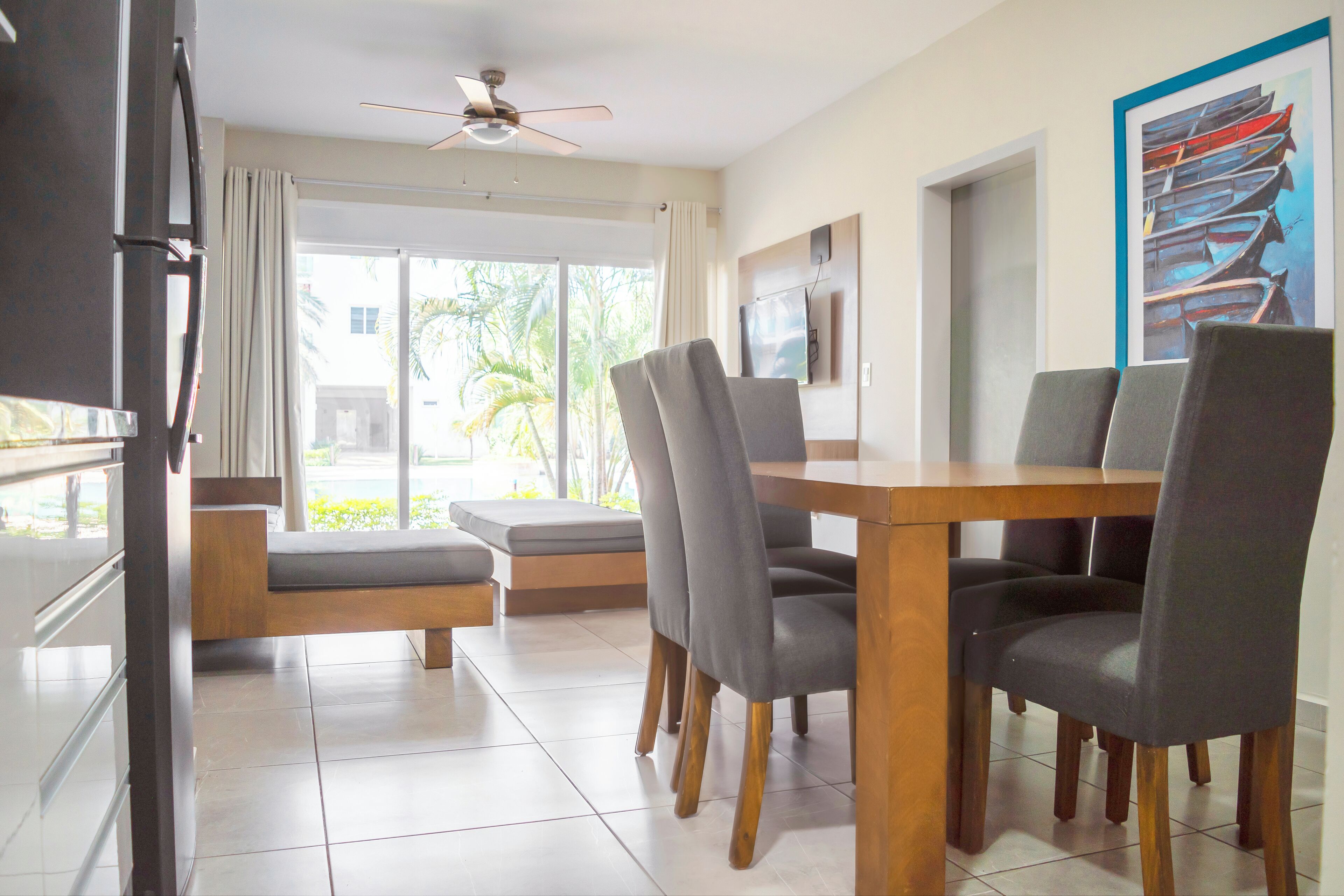 A modern dining area with a wooden table and grey upholstered chairs, a ceiling fan, and a large window with curtains.