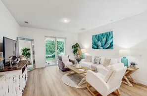 A spacious living room with a white sofa, a coffee table, and a TV on a wooden cabinet. There is a large window with a view of greenery outside. - Boaters Cove ~ 3 Car Garage Home (Sebastian)
