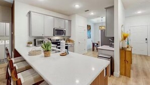 A modern kitchen with white cabinetry, a central island, and a dining area with a chair and table. - Boaters Cove ~ 3 Car Garage Home (Sebastian)