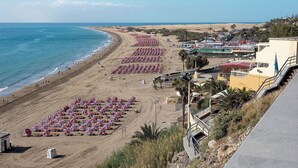 A beach with rows of umbrellas and sun loungers, a coastal town in the background, and a clear sky. - Tamaran (San Bartolomé de Tirajana)