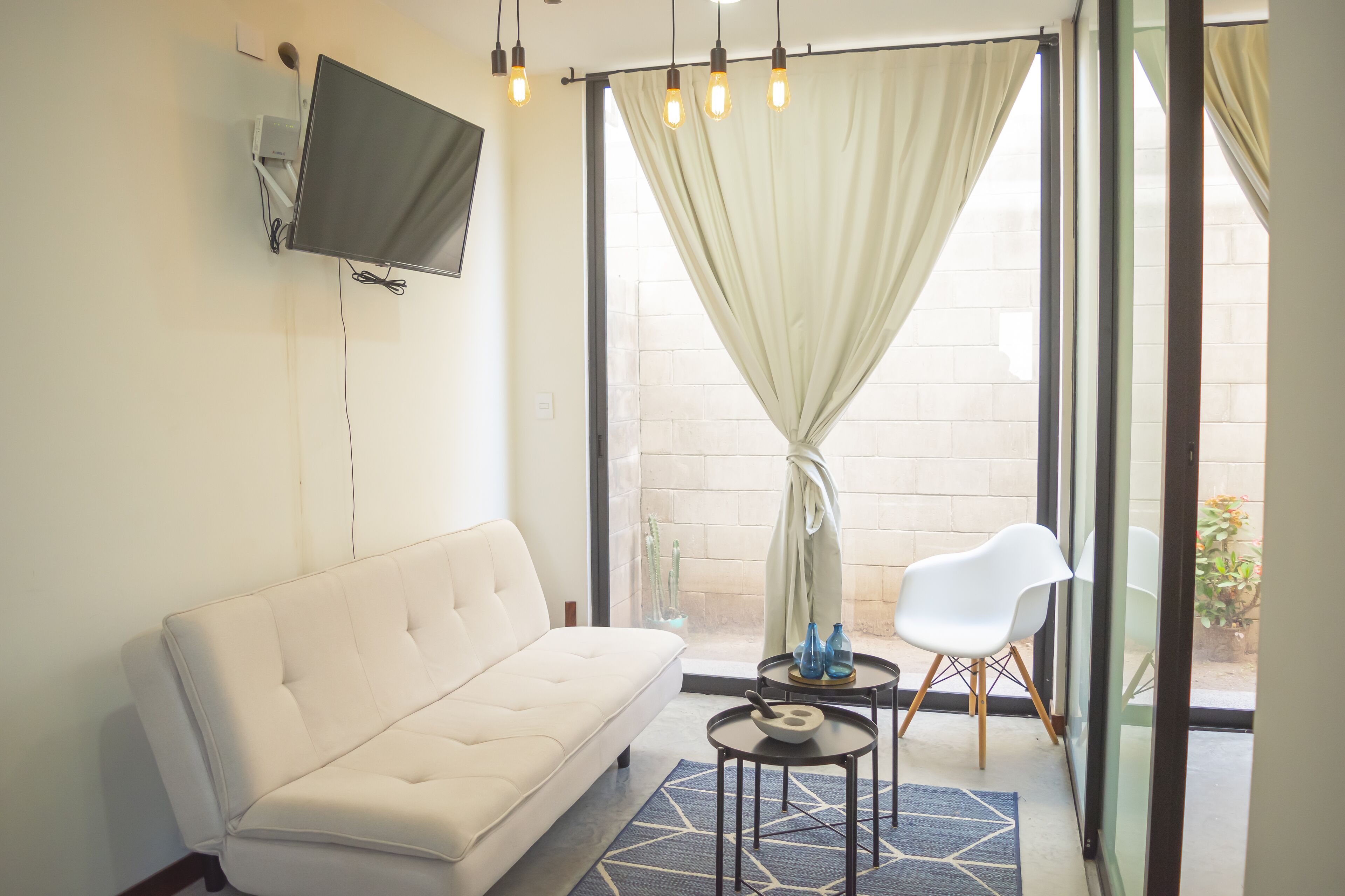 A modern living room with a sofa, a flat-screen TV, a small table with decorative items, and a view through large windows.