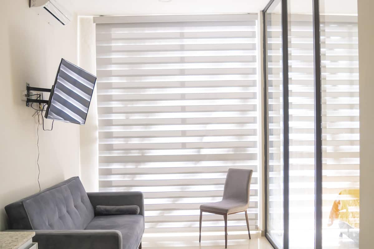 A modern living room with a sofa, a chair, a flat-screen TV, and a large window with blinds.