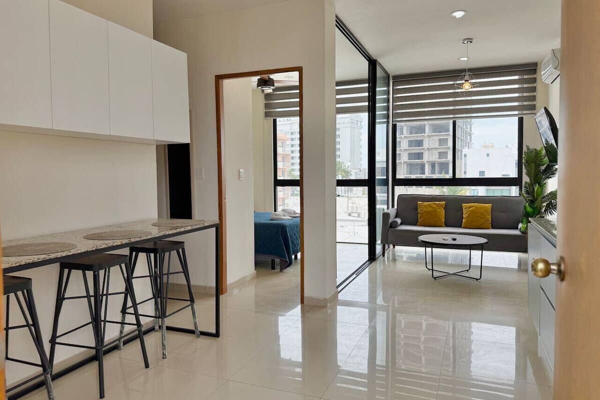 A modern kitchen with a bar area, a dining table, and a sofa in a bright living area with large windows.