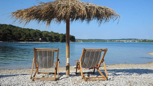 Two wooden beach chairs with striped cushions under a thatched umbrella on a pebbled beach with a calm sea and a distant shoreline. - Charming 2-Bedroom Haven for Four, No Pets (Pula)