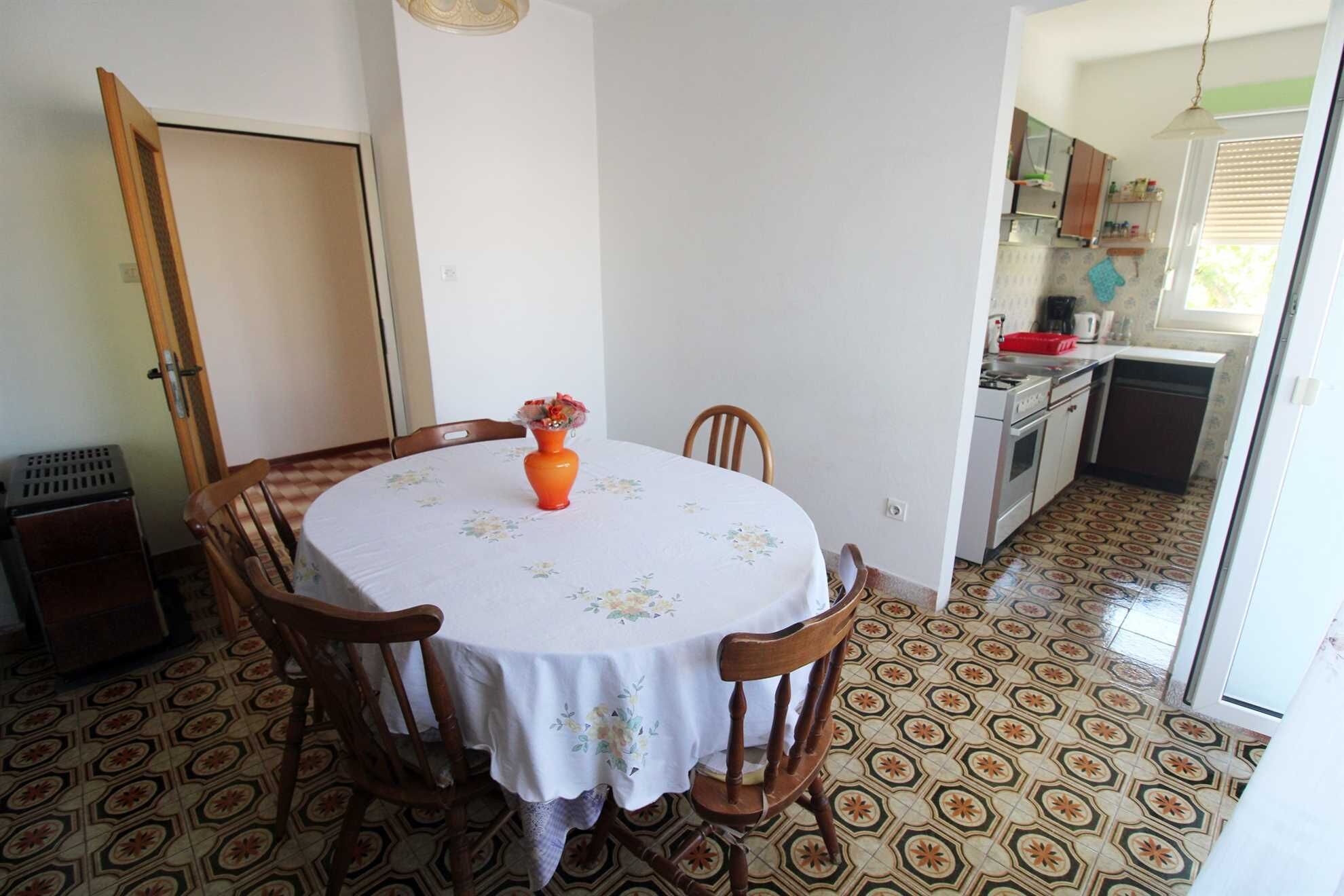 A dining area with a round table, chairs, a vase with flowers, and a kitchen in the background.
