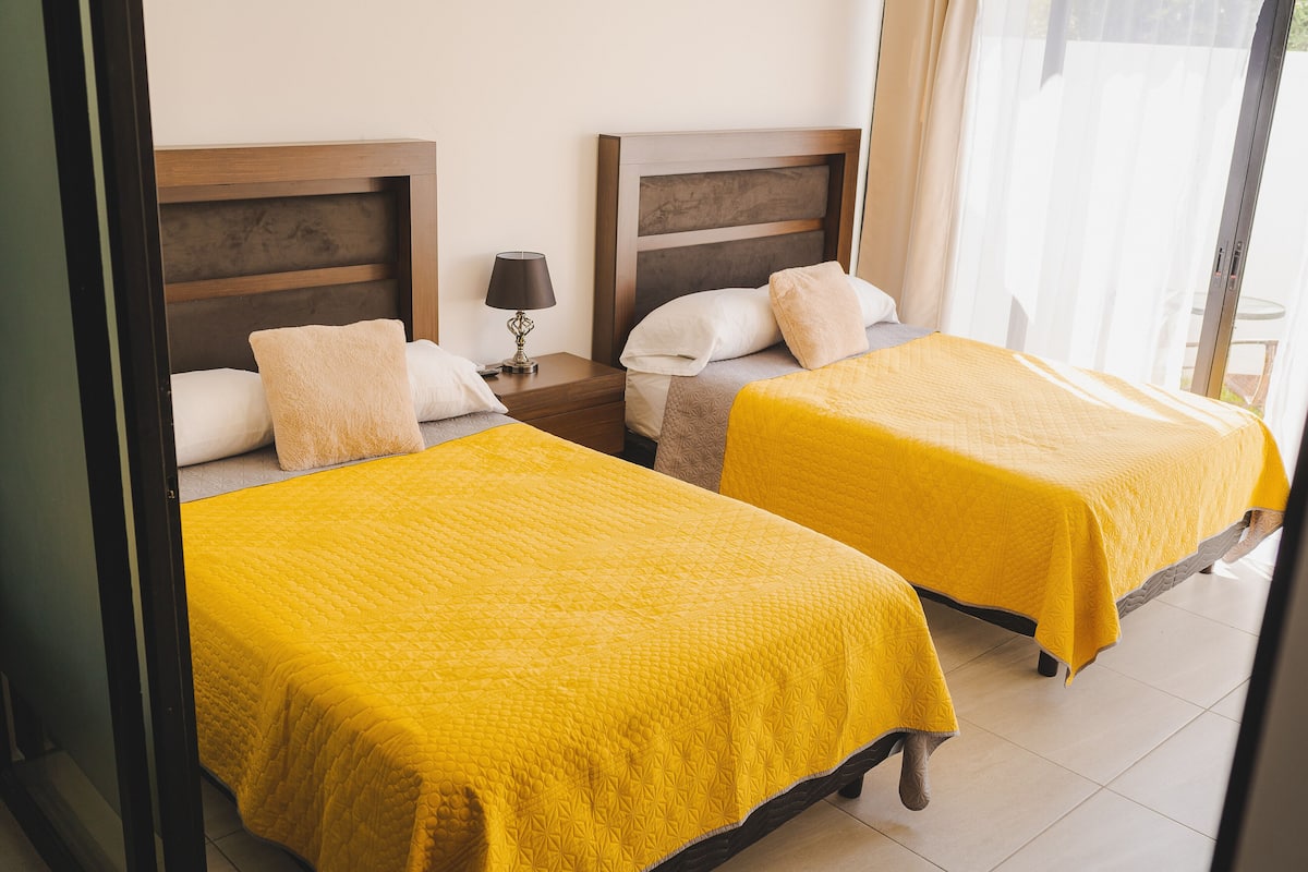 A hotel room with two single beds, each with a yellow bedspread, wooden headboards, and a nightstand with a lamp.