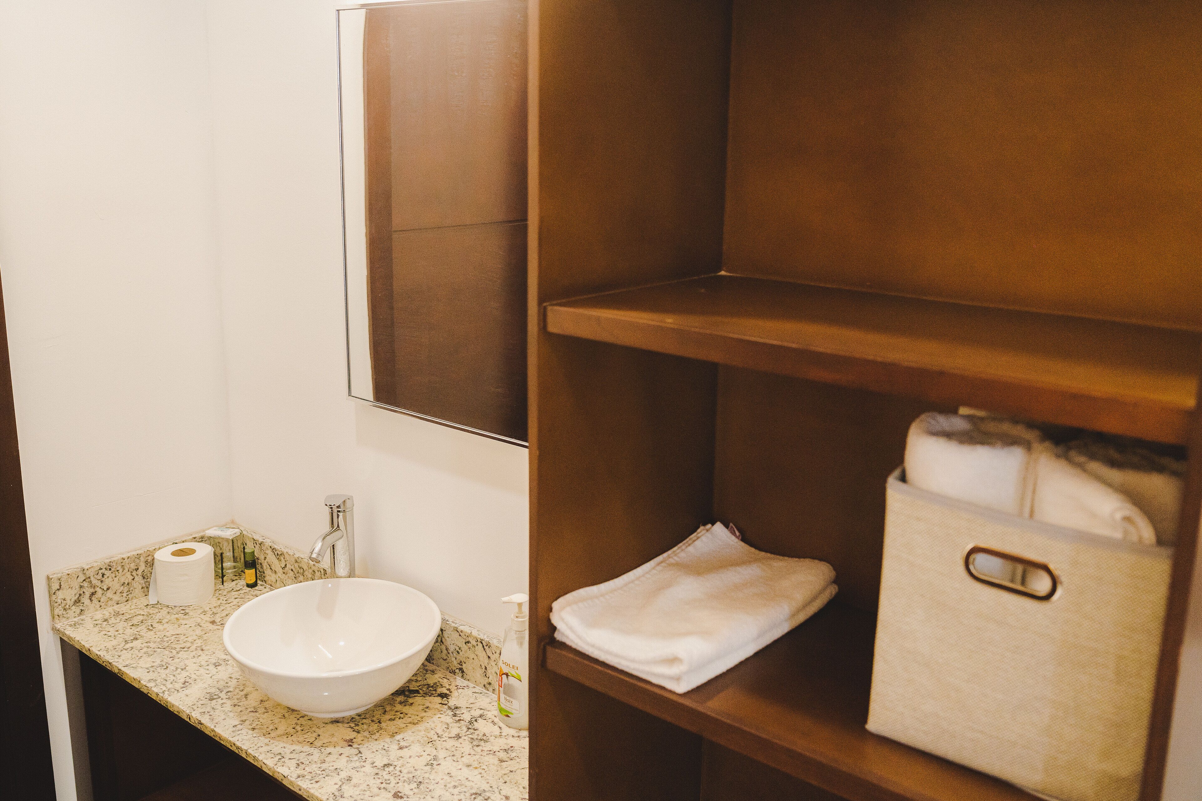 Un baño con encimera de mármol, un lavamanos blanco, un espejo y un estante de madera con toallas y artículos de aseo.