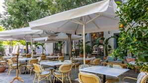 Café al aire libre con sillas y mesas de mimbre, cubierto por sombrillas blancas, rodeado de plantas y árboles en macetas.