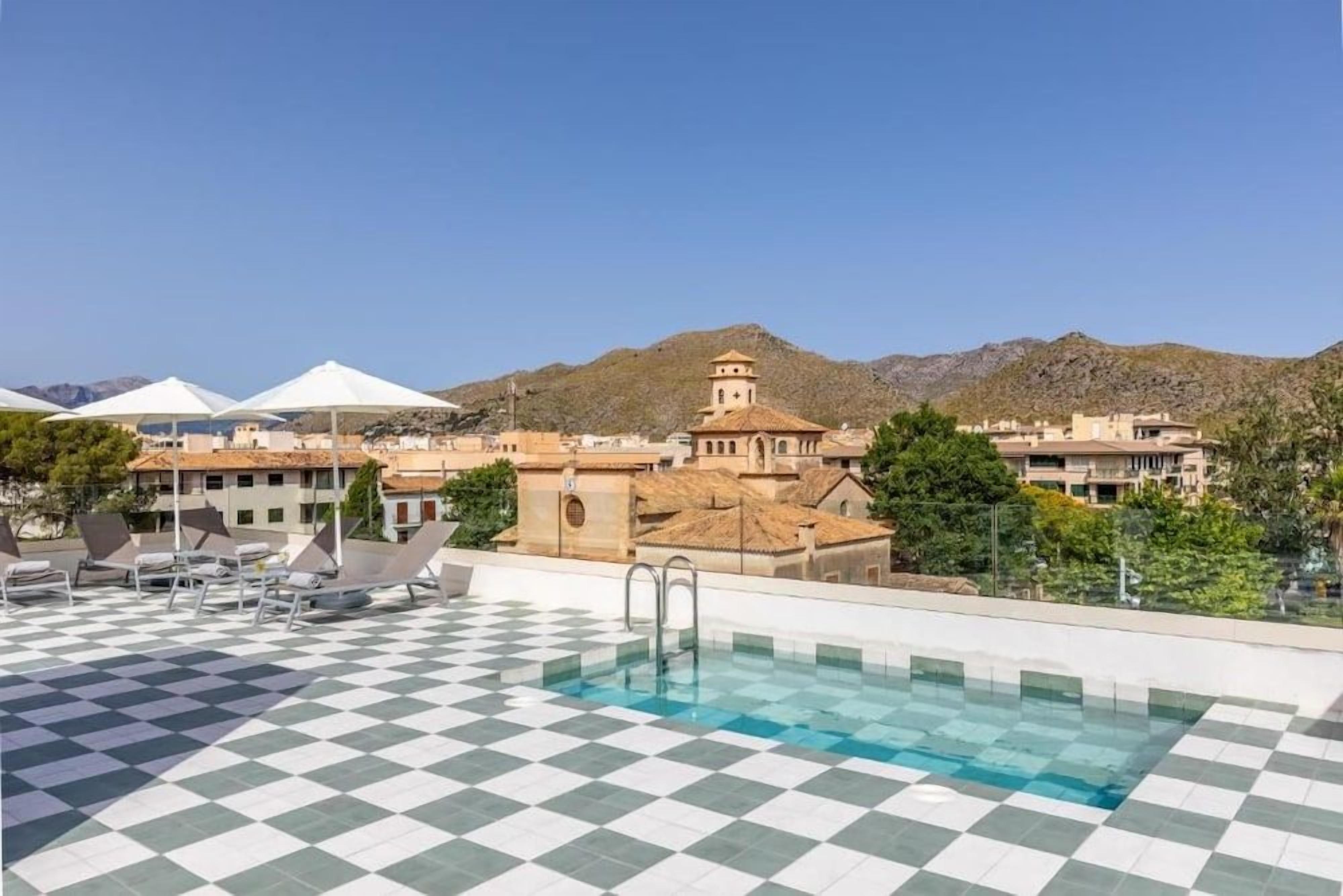 A rooftop pool with a checkered floor, lounge chairs, and a view of buildings and mountains in the background.