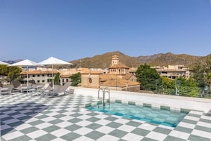 Una piscina en la azotea con suelo a cuadros, sillones de descanso y vistas a edificios y montañas al fondo.