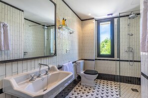 Um banheiro moderno com pia grande, vaso sanitário e chuveiro. Possui um espelho grande, uma janela e piso revestido de azulejos.