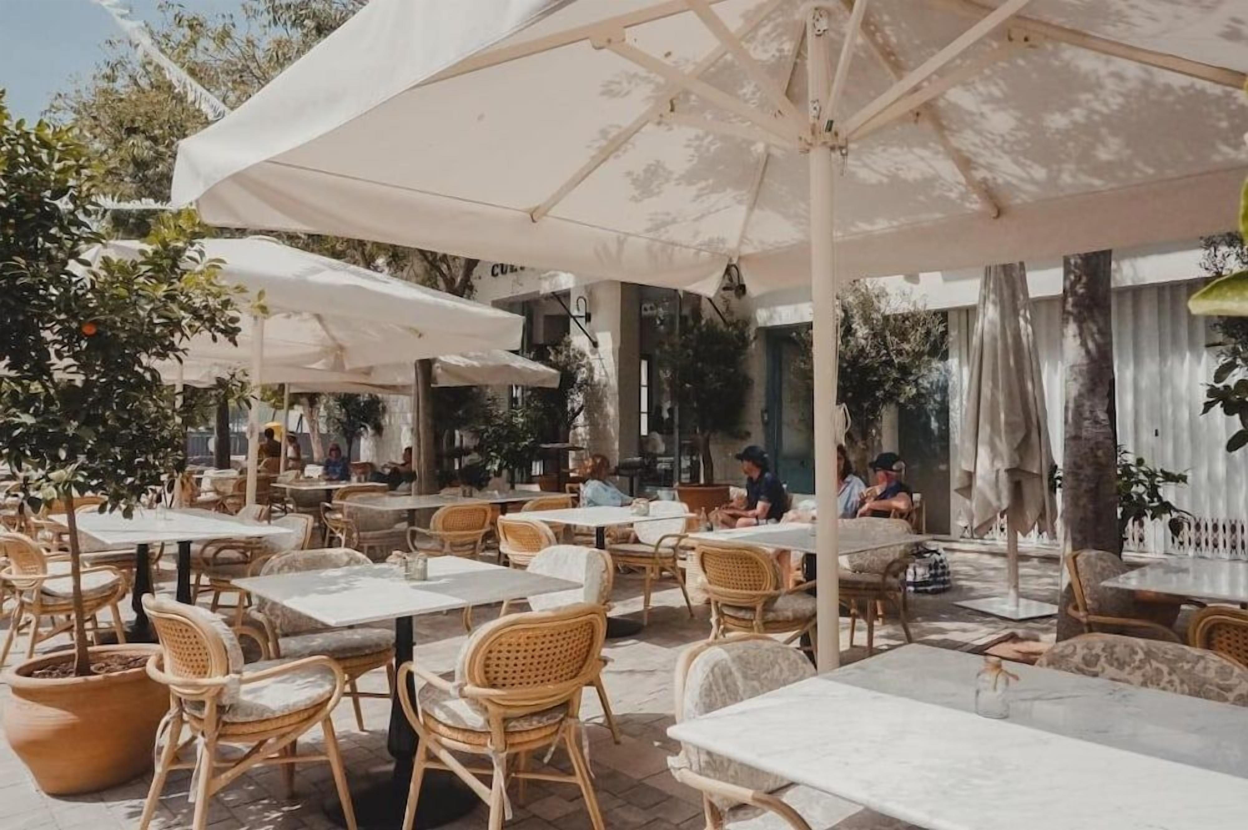 An outdoor cafe with tables and chairs under large umbrellas, surrounded by trees and buildings.