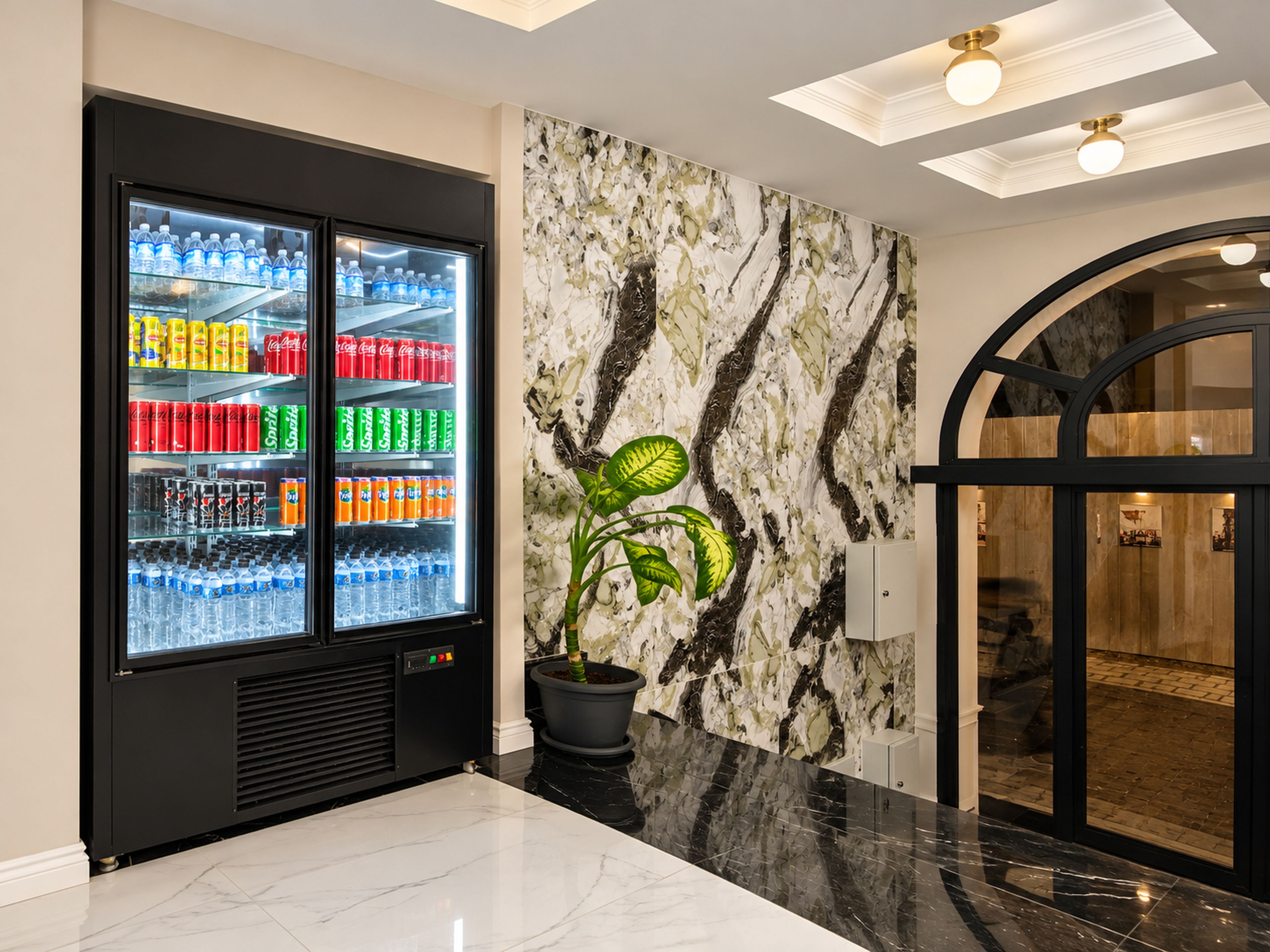 A modern lobby with a beverage refrigerator stocked with various drinks, a marble-patterned wall, and a potted plant.